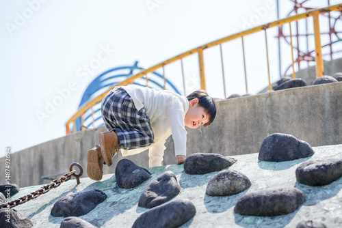 In spring at a park in Fukuoka, a 3-year-old Japanese boy climbs a rock playground structure. This photo captures his spirit of adventure and joy as he explores the play area on a sunny spring day.