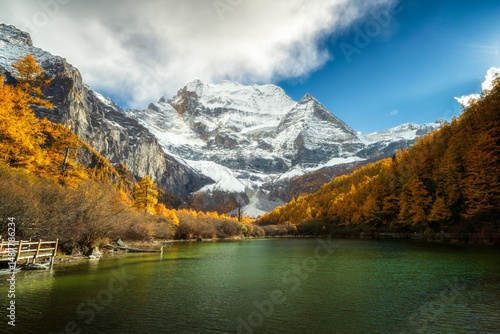 Snow capped mountain, autumn forest, clear lake, blue sky, and wooden walkway create peaceful landscape with vibrant colors and natural beauty