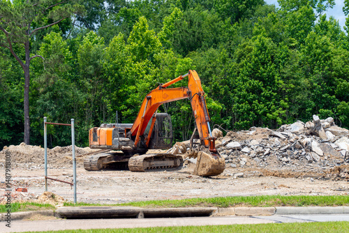 An orange and black backhoe is actively engaged at a construction site, clearing a pile of rubble composed of concrete and debris. The machinery is equipped with a hydraulic arm and bucket for digging