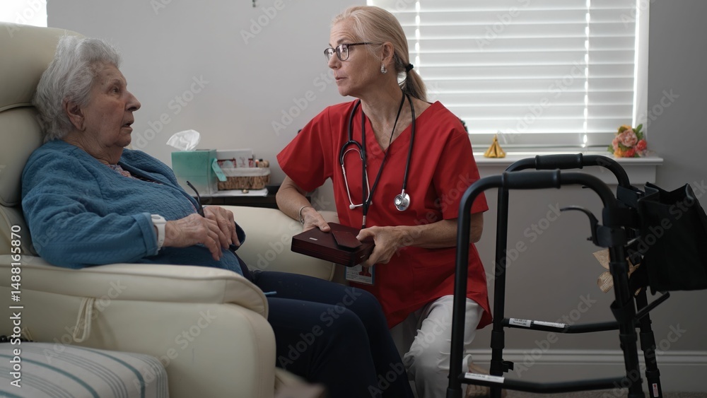 Obraz premium A healthcare worker in a red uniform engages with an elderly woman sitting in a comfortable chair, fostering a caring atmosphere during a home visit.