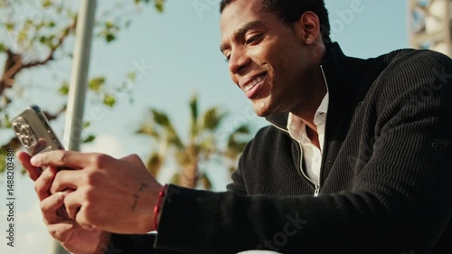 A young man sits outdoors on a sunny day, looking at his phone and smiling. He appears happy and engaged with the content on his device.