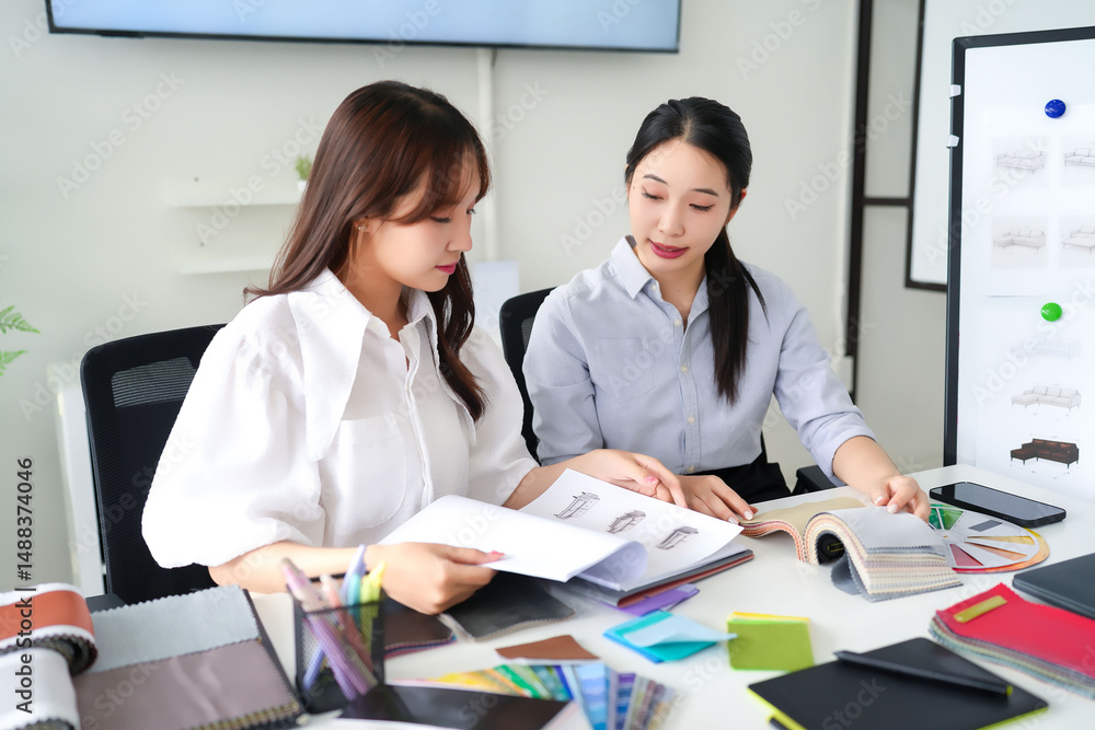 Obraz premium Two Asian Interior Designers Collaborating on Fabric and Sofa Designs in a Modern Office Young Women Discussing Home Decor and Furniture Choices for Clients