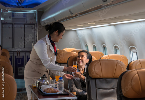 Cheerful flight attendant serving orange juice drink to smiling female passenger in airplane cabin. In flight service cart with  teapot cups water bottles and pastry