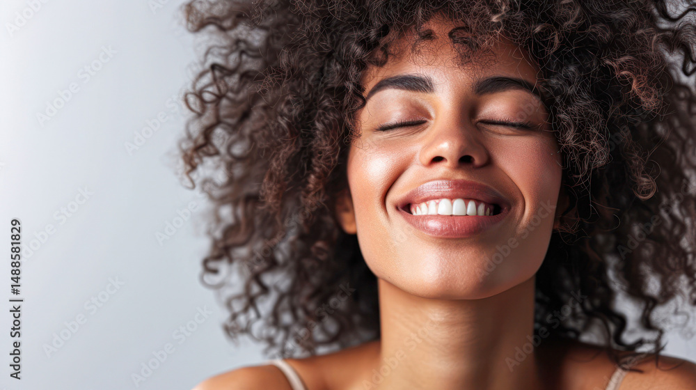 A woman with curly hair smiles joyfully with her eyes closed, radiating happiness and confidence against a light background. A woman with curly hair smiles joyfully with her eyes closed, radiating happiness and confidence against a light background.