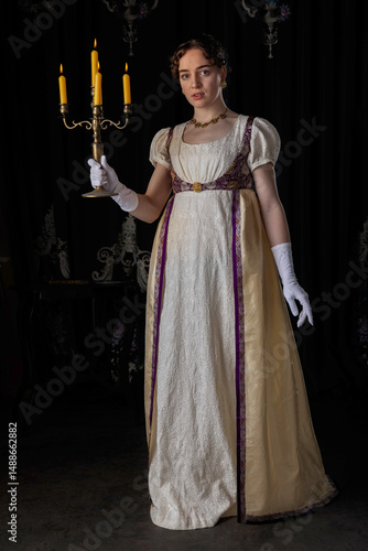 A Regency woman wearing evening attire, standing and holding a lit candelabra
