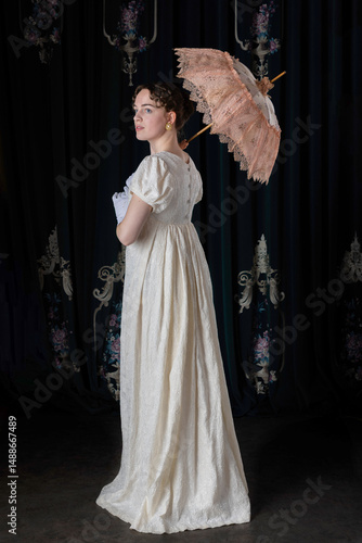 A Regency woman wearing an embroidered dress and holding a parasol, standing in front of velvet curtains