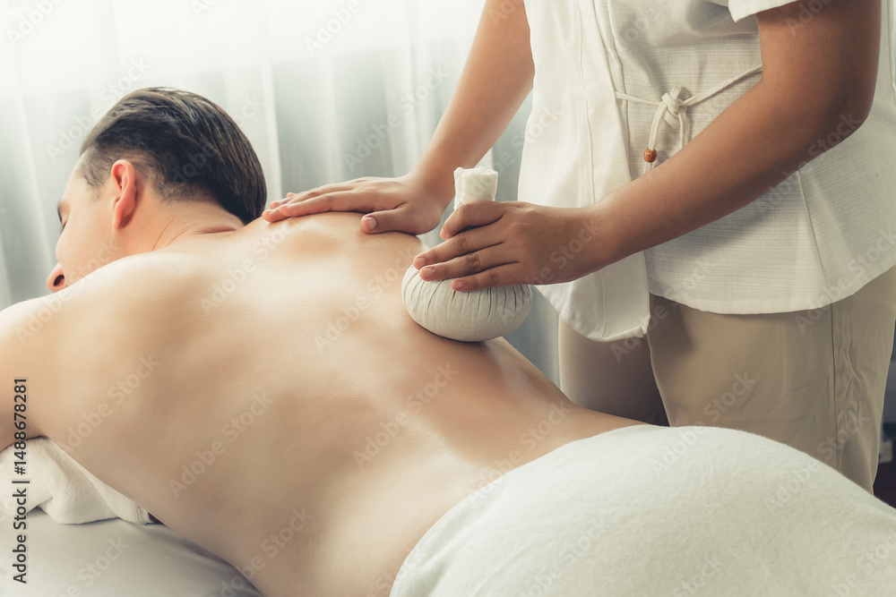 Obraz premium Hot herbal ball spa massage body treatment, masseur gently compresses herb bag on man body. Tranquil and serenity of aromatherapy recreation in day lighting ambient at spa salon. Quiescent