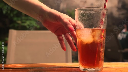A hand reaches for a cold glass of iced lemon tea covered with condensation. The perfect refreshing summer drink to quench thirst in hot weather with bright citrus and cool moisture.