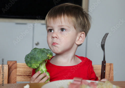 portrait of cute toddler sitting at the table with plate with noodles and sausages holding broccoli  with a face of disgust  