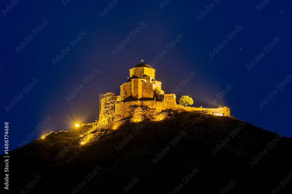 Obraz premium Night view of Jvari Monastery. Stone walls are illuminated with warm light. Traces of spotlights are visible. Dark blue sky with stars in the background