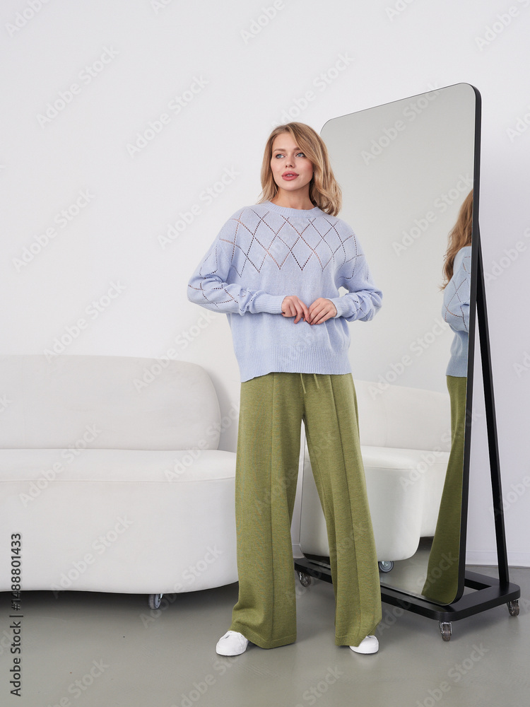 Ταπετσαρία A woman in a light blue sweater and green pants stands near a floor mirror, reflecting her image