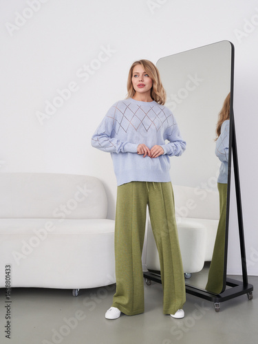 Tableau sur toile A woman in a light blue sweater and green pants stands near a floor mirror, reflecting her image