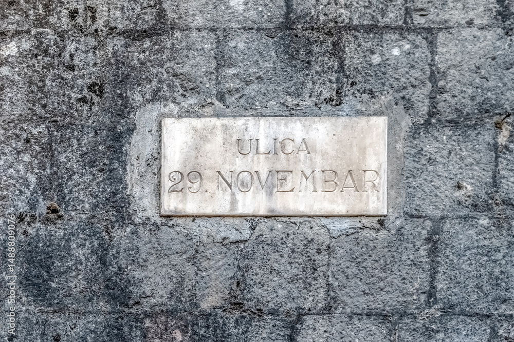 Fototapeta premium Street sign Ulica 29. Novembar on a stone wall in Kotor. The sign commemorates a historic date, reflecting the cultural and historical significance of the area. Kotor, Montenegro - June 18, 2021