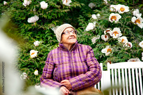 An old woman in a park on a bench with a tree peony flower. An old lady in a boarding house enjoys life. The concept of care, old age and happiness. 