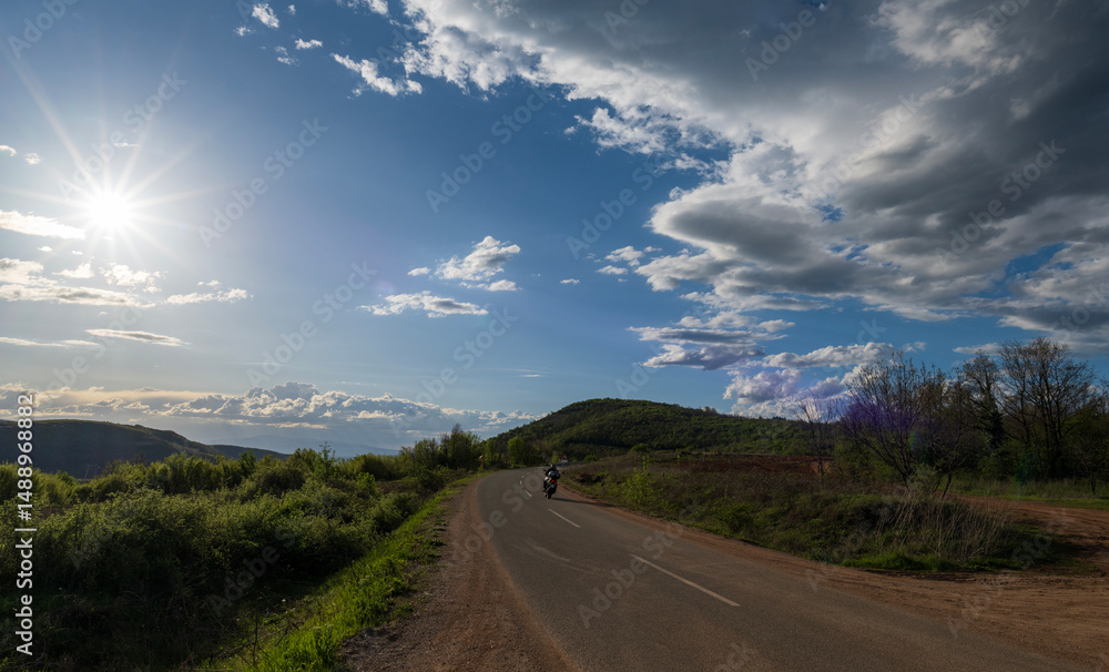 Fototapeta premium A man on a motorcycle drives on a highway that passes through hills and mountains. The sunset, white clouds and blue sky can be seen in the background. 
