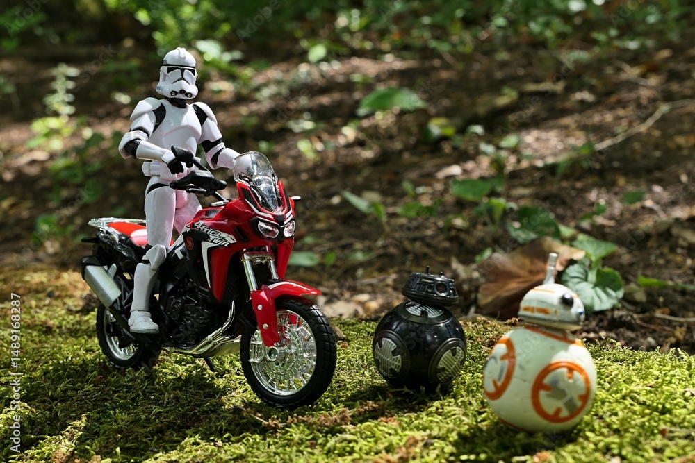 Naklejka premium Star Wars action figure of Clone Trooper, riding on Honda Africa Twin red dual sport motorbike, two ball shaped astromech droids, imperial BB-9E and BB-8 rolling on forest moss in front of him. 