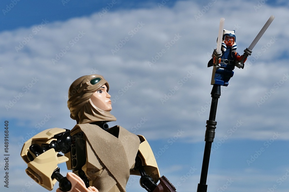 Fototapeta premium LEGO Star Wars action figure of female Jedi Rey Skywalker looking at small figure of female Togruta Jedi Ahsoka Tano with two lightsabers on tip of her pole. Cloudy skies background. 