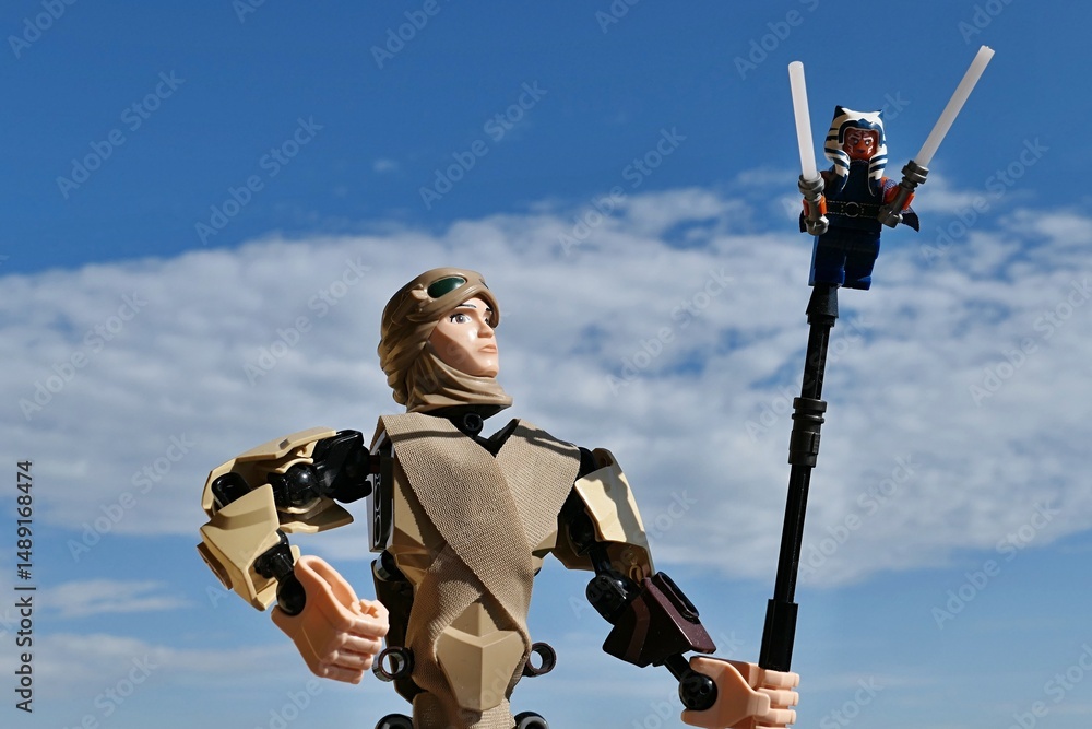 Fototapeta premium LEGO Star Wars action figure of female Jedi Rey Skywalker holding small figure of female Togruta Jedi Ahsoka Tano with two lightsabers on tip of her pole. Cloudy skies background.