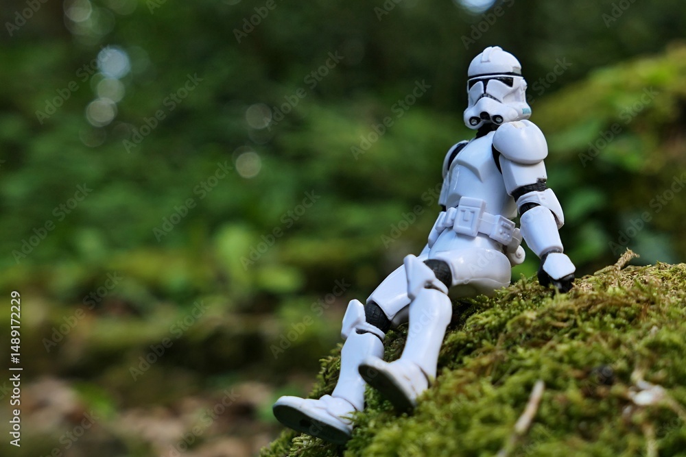 Fototapeta premium Star Wars articulated action figure of Clone Trooper soldier in white armor, sitting relaxed on mossy stone in shadow of spring foliage. 