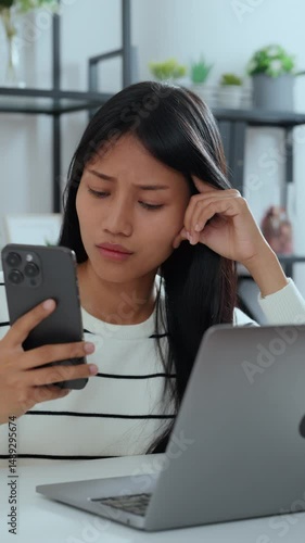 Young asian woman feeling stressed and anxious while looking at her smartphone, sitting at desk with laptop, concept of online problem, finance issue, or digital stress while working remotely
