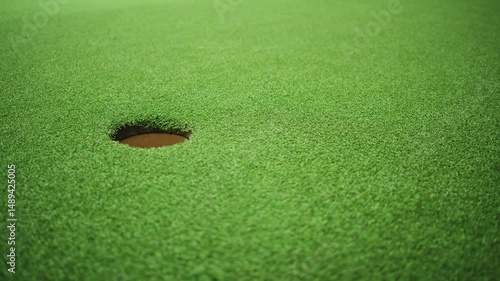 Golf ball rolling toward hole on green turf in wide frame slow motion