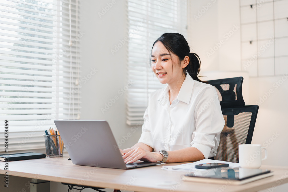 Young Asian woman working at desk with laptop, smiling in bright modern office, natural light from window, coffee cup and stationery on table, professional and productive atmosphere Young Asian woman working at desk with laptop, smiling in bright modern office, natural light from window, coffee cup and stationery on table, professional and productive atmosphere