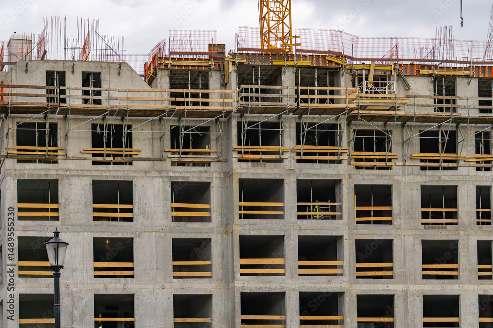 View of a multi-story building under construction, with visible window openings, formwork, and scaffolding. The concrete structure is in the raw state, and a portion of a tower crane is visible at the View of a multi-story building under construction, with visible window openings, formwork, and scaffolding. The concrete structure is in the raw state, and a portion of a tower crane is visible at the