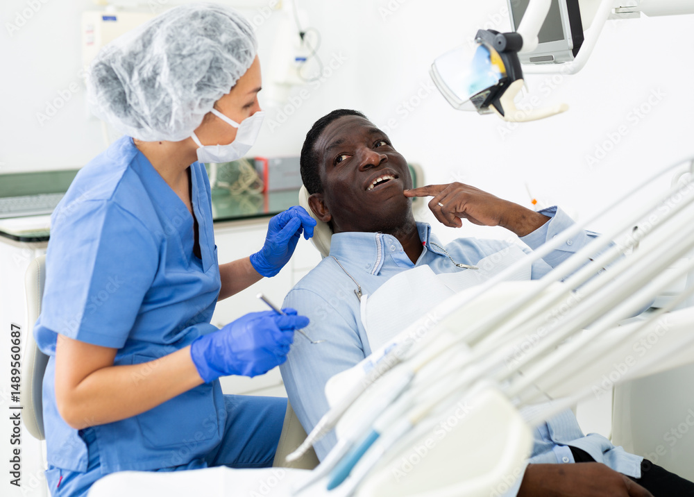 Obraz premium African-american man pointing at his teeth during consultation, woman dentist sitting and listening to him.
