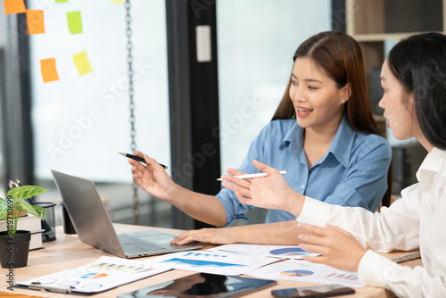 Foto Two Asian businesswomen holding graph papers are sitting and talking happily while working