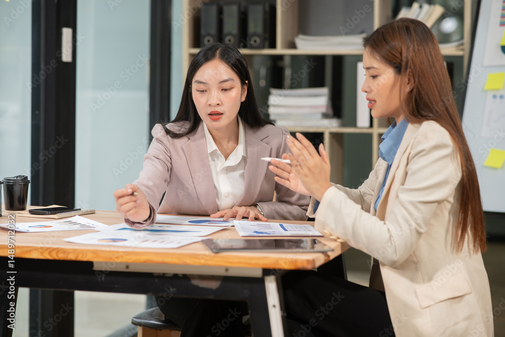 Two Asian businesswomen holding graph papers are sitting and talking happily while working.
Two Asian businesswomen holding graph papers are sitting and talking happily while working.