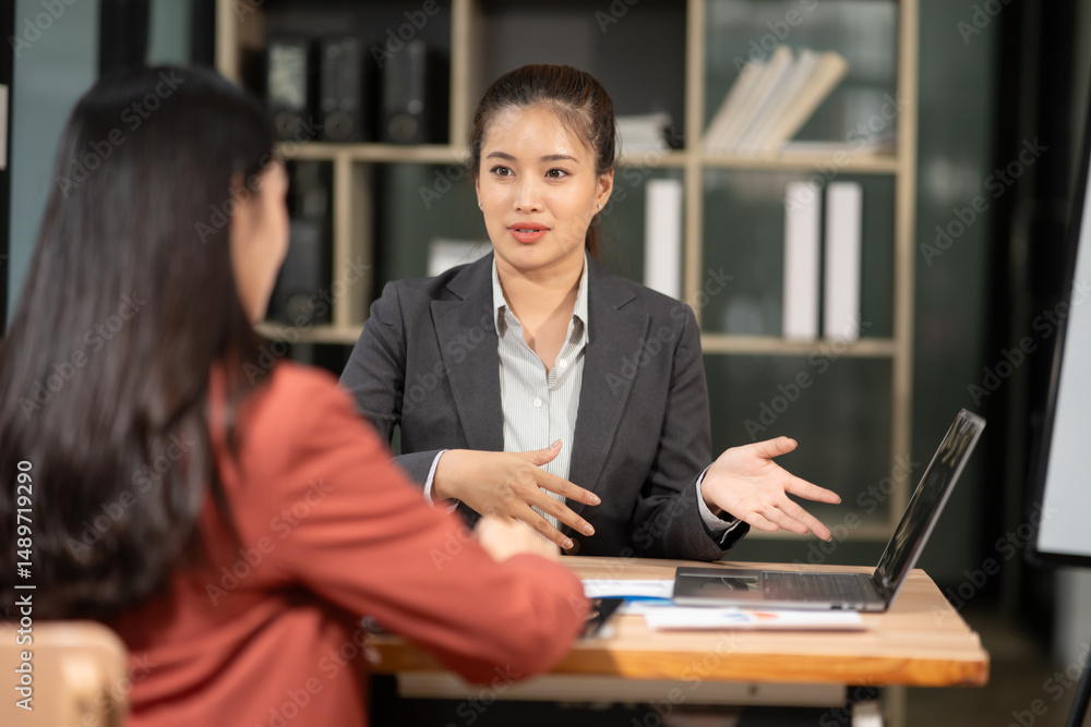 Obraz premium Two Asian businesswomen with laptops discussing documents in an office.