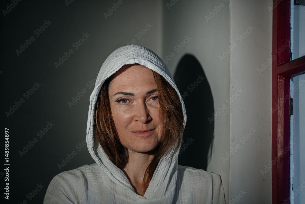 Caucasian female in hooded attire standing by window in shadowed room Caucasian female in hooded attire standing by window in shadowed room