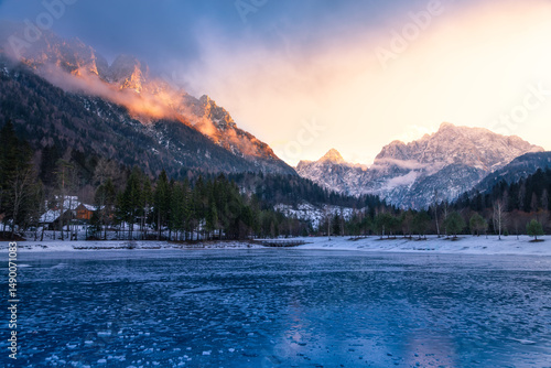 Frozen lake Jasna in Kranjska Gora (Slovenia) reflects snow-dusted mountains under a dramatic, colorful sky at dawn or dusk.