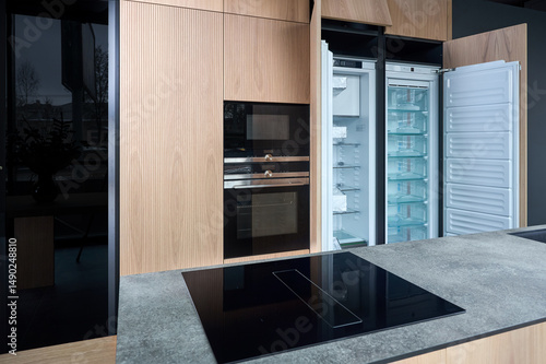 Contemporary fitted kitchen interior with minimalist flat design with wooden striped cabinets, built-in oven microwave and empty fridge with opened doors and island with hob and stoneware countertop.
