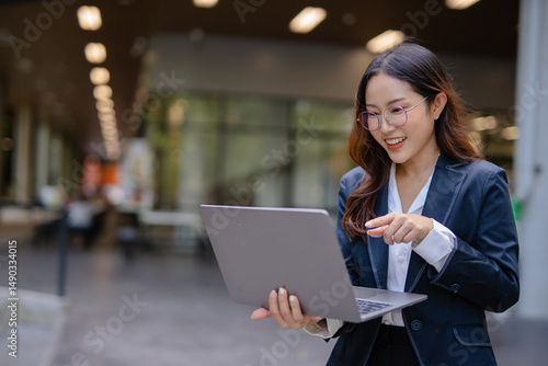 Asian businesswoman using digital laptop while standing in front of modern building. Attractive female employee working in front of office. Free Download Asian businesswoman using digital laptop while standing in front of modern building. Attractive female employee working in front of office. - GFXLoad
