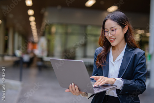 Asian businesswoman using digital laptop while standing in front of modern building. Attractive female employee working in front of office.