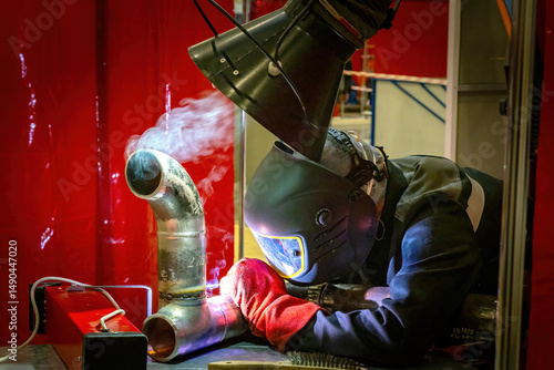 Welder in full protective gear working on metal pipe with visible smoke under extraction hood in workshop. Metal fabrication. Pipe welding. Industrial safety.