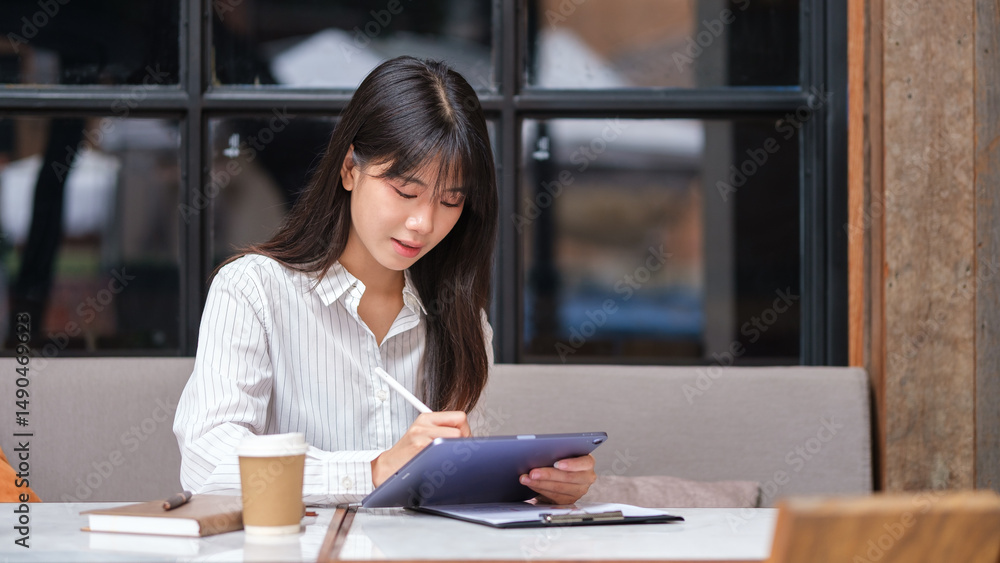 Fototapeta premium Professional Asian woman planning or working on a tablet with coffee and notes beside her.