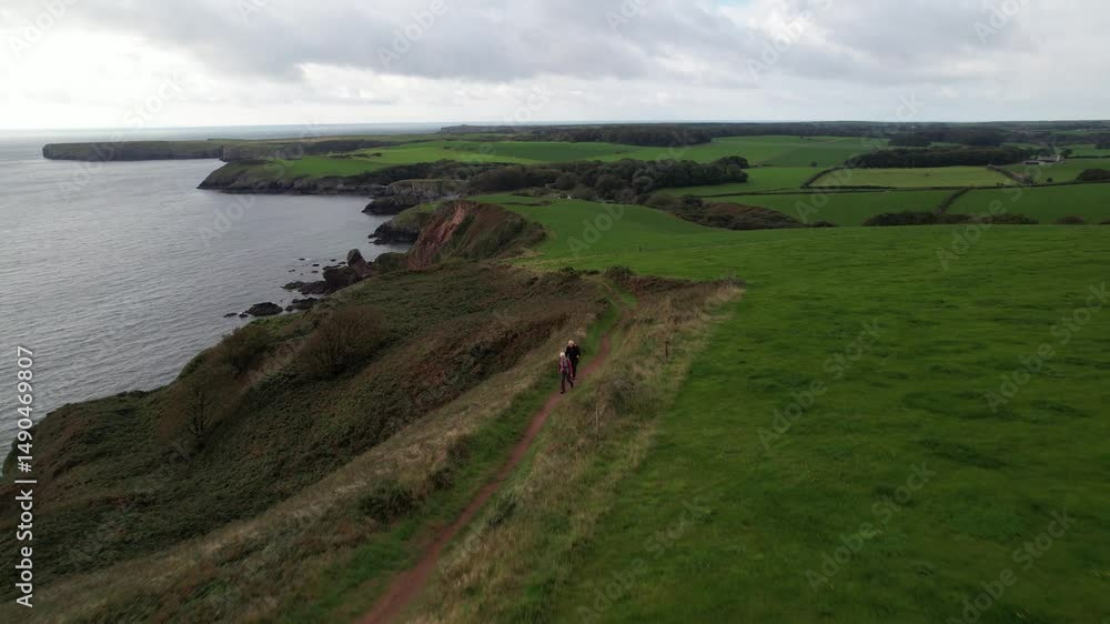 green meadows and rugged rocky cliffs at sea coast. Backwards reveal of hikers walking on pathway. South Wales, UK green meadows and rugged rocky cliffs at sea coast. Backwards reveal of hikers walking on pathway. South Wales, UK