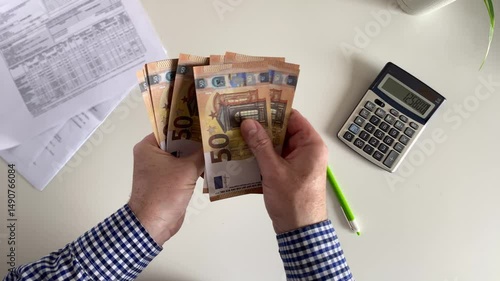A man counts 50 euro paper bills against the background of utility bills and a calculator.