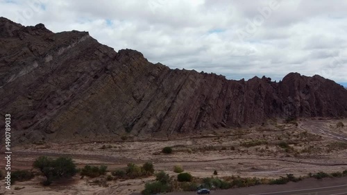 Stunning aerial shot approaching the Cañadon del Indio in Fiambala, Catamarca. Mountains, gorges, the almost dry river, cloudy sky, the Route of the 6 Miles, and vehicles at the viewpoint are visible.