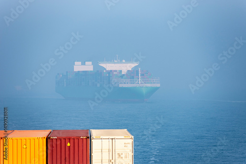 Фотография A large cargo ship laden with containers is partially obscured by fog, sailing on a calm sea
