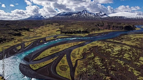 Spectacular aerial view of a meandering river flowing through a rugged landscape, with mountains in the background. The river's striking blue color contrasts with the surrounding dark terrain