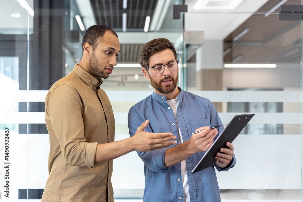 Two business professionals discuss paperwork, one pointing and the other holding a pen, in a modern office setting. Two business professionals discuss paperwork, one pointing and the other holding a pen, in a modern office setting.