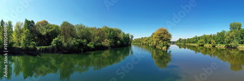 Die Lechmündung in die Donau zeigt eine friedliche Flusslandschaft mit üppiger Vegetation und klarem Wasser. Die Spiegelung der Bäume im Wasser und der blaue Himmel schaffen eine idyllische Atmosphäre