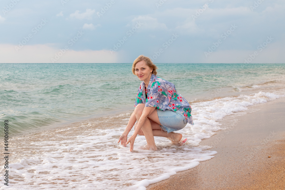Woman in floral shirt and denim shorts squatting playing in shallow waves on sandy beach. Summer vacation at sea Woman in floral shirt and denim shorts squatting playing in shallow waves on sandy beach. Summer vacation at sea
