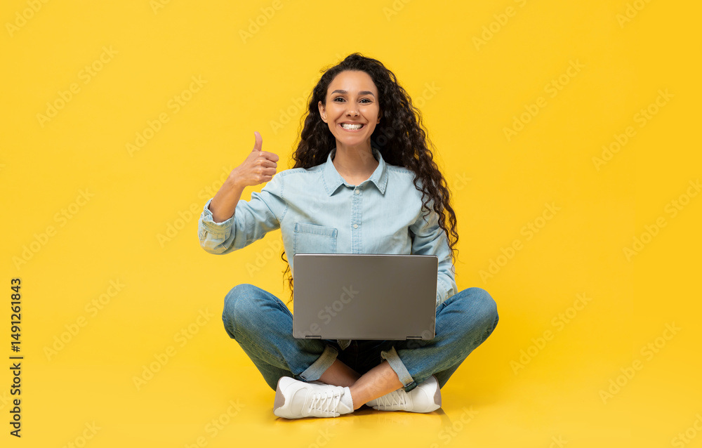 Naklejka premium I Like. Portrait Of Happy Excited Young Woman Sitting On Floor At Studio Using Pc Laptop And Showing Thumbs Up Sign Gesture, Working Or Studying Remotely, Posing To Camera, Yellow Orange Wall