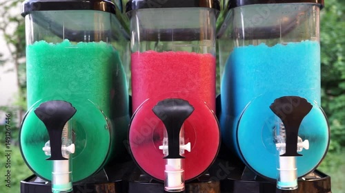 Three colorful frozen drink containers filled with slushy ice in green, pink, and blue, ready for dispensing on a hot day.