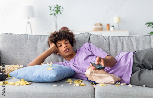 Lazy black teen guy lying on sofa with scattered food, holding TV controller, watching dull movie on television at home. Bored African American adolescent killing time indoors during covid lockdown
