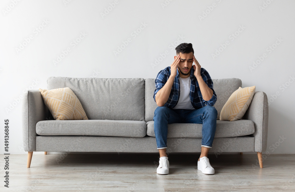 Fototapeta premium Sad young Middle Eastern man suffering from headache or migraine, touching head massaging temples. Stressed guy sitting on couch with painful face expression feeling terrible weakness or depression